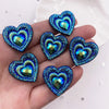 A hand holds a set of twelve heart-shaped blue and green iridescent peacock resin rhinestone flatbacks.