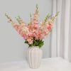 Un vase rempli de fleurs artificielles de delphinium roses et blanches, exposé contre un mur gris clair.
