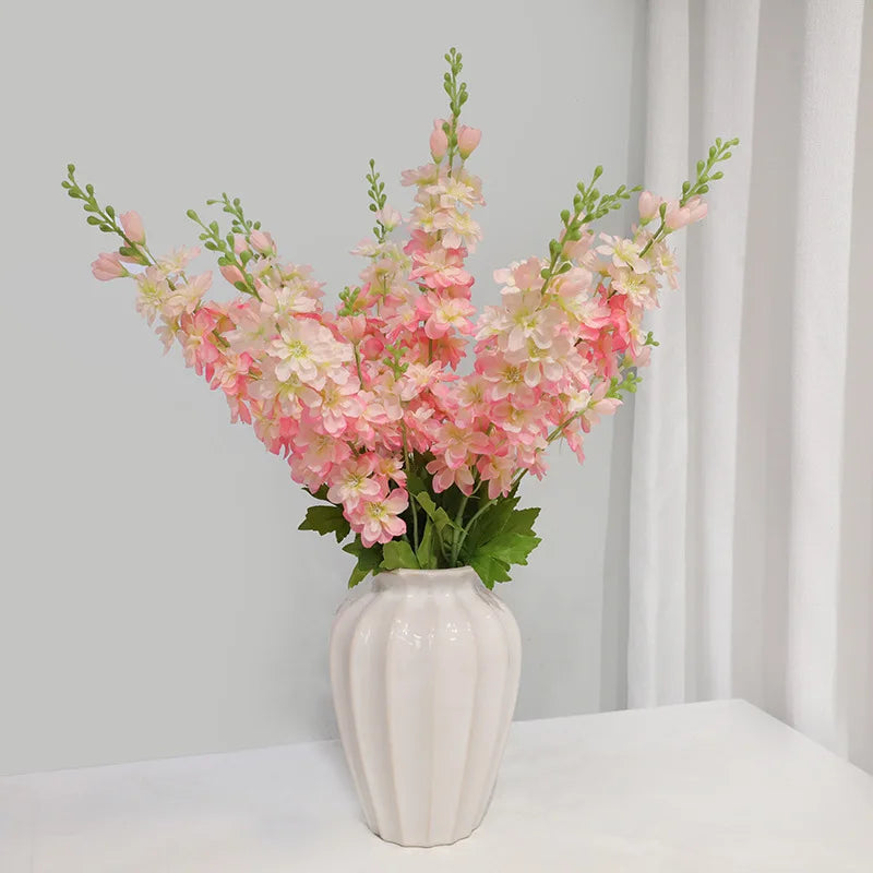 Un vase rempli de fleurs artificielles de delphinium roses et blanches, exposé contre un mur gris clair.