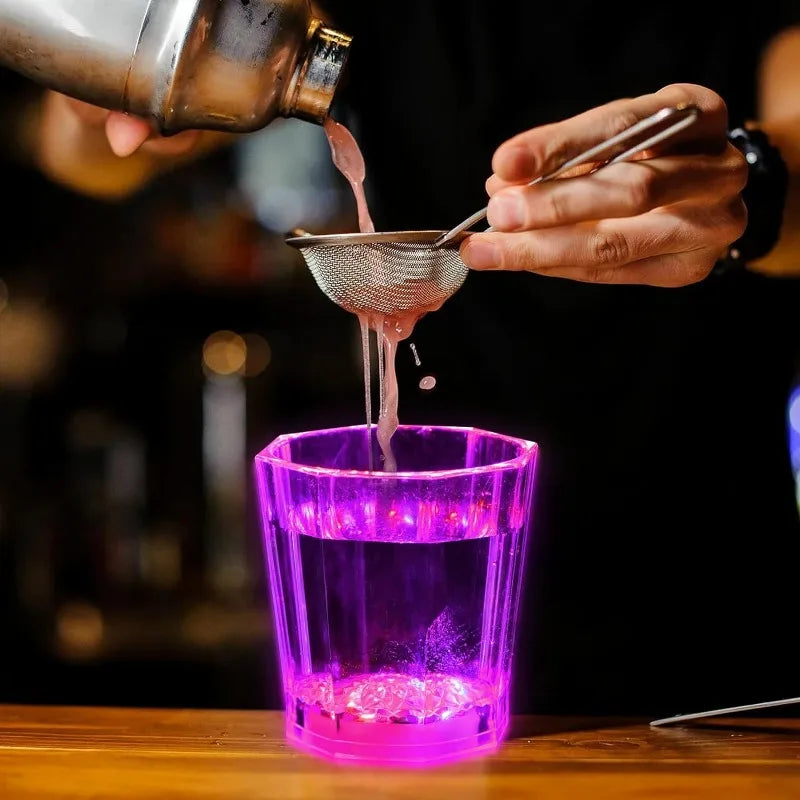 A hand pours a liquid from a bottle through a sieve into a glowing purple shot glass.