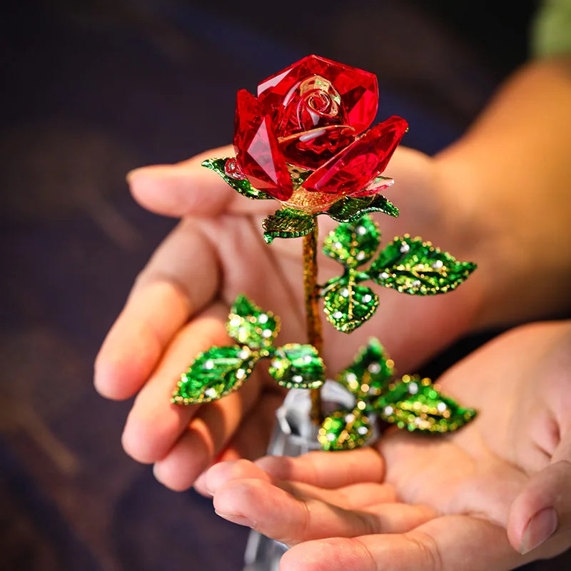 A hand holds a handcrafted crystal glass rose vase with a red rose and golden leaves.