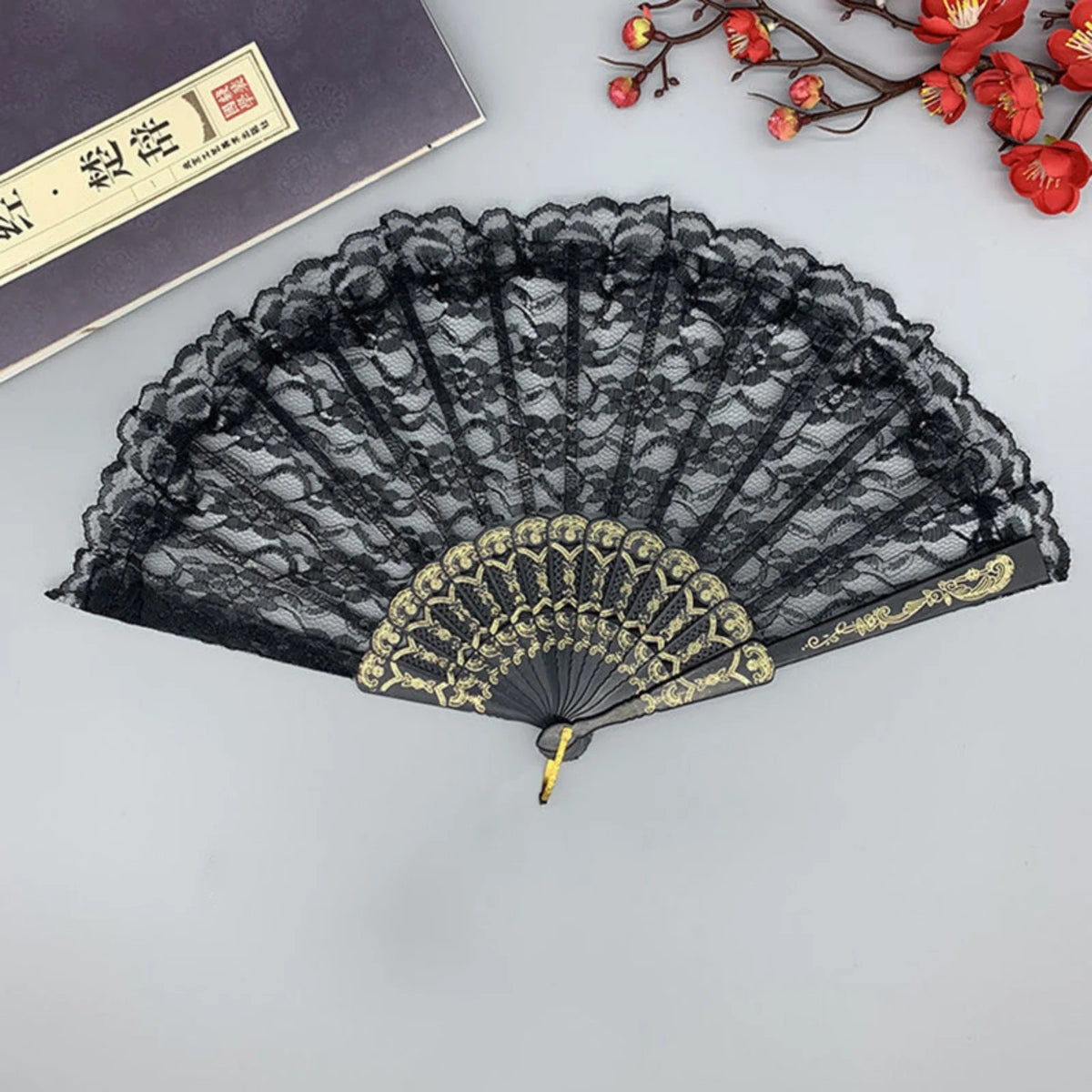 Abanico de mano plegable de encaje vintage negro con detalles dorados, exhibido sobre una superficie gris.