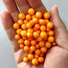 A hand holds a pile of small, round, orange foam beads for crafting and decoration.