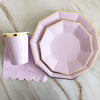 A pink and gold party tableware set featuring a plate and a cup resting on a purple napkin on a marble surface.