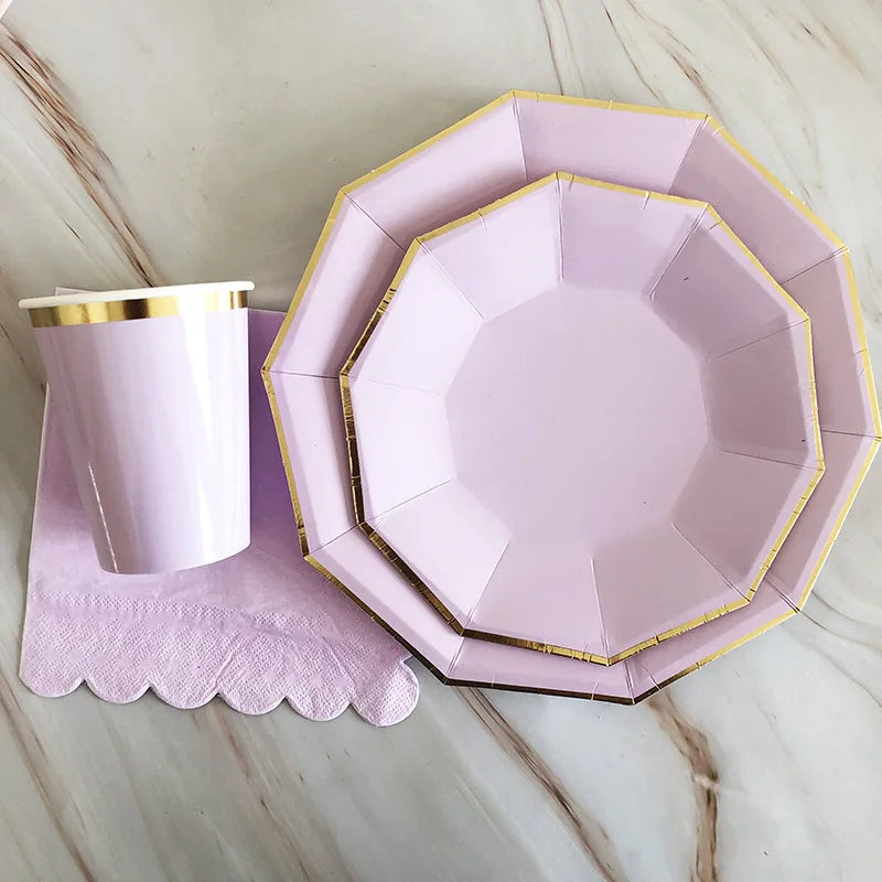 A pink and gold party tableware set featuring a plate and a cup resting on a purple napkin on a marble surface.