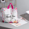 White floral thank you gift bags with pink handles and tissue paper displayed on a gray surface.