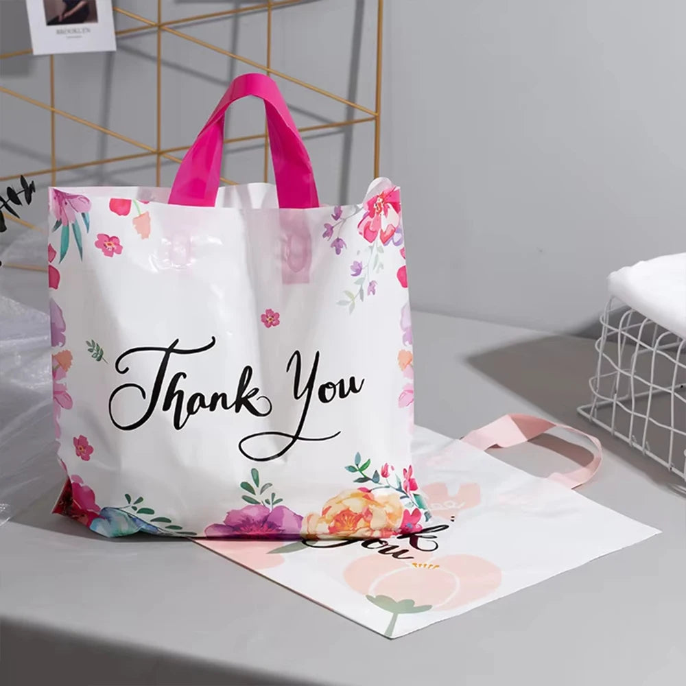 White floral thank you gift bags with pink handles and tissue paper displayed on a gray surface.