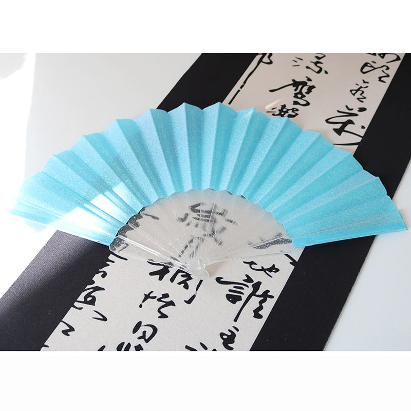 A light blue glitter folding fan with transparent ribs is displayed against a black background with Japanese calligraphy.