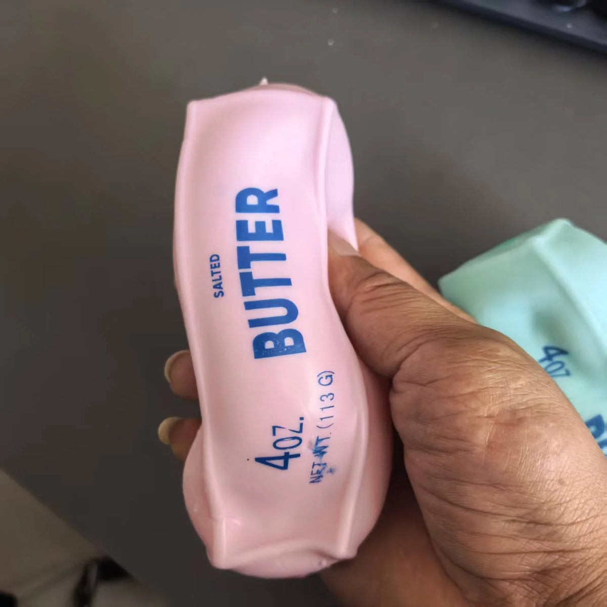 A hand holds a pink butter squeeze toy labeled 4oz salted butter for stress relief.