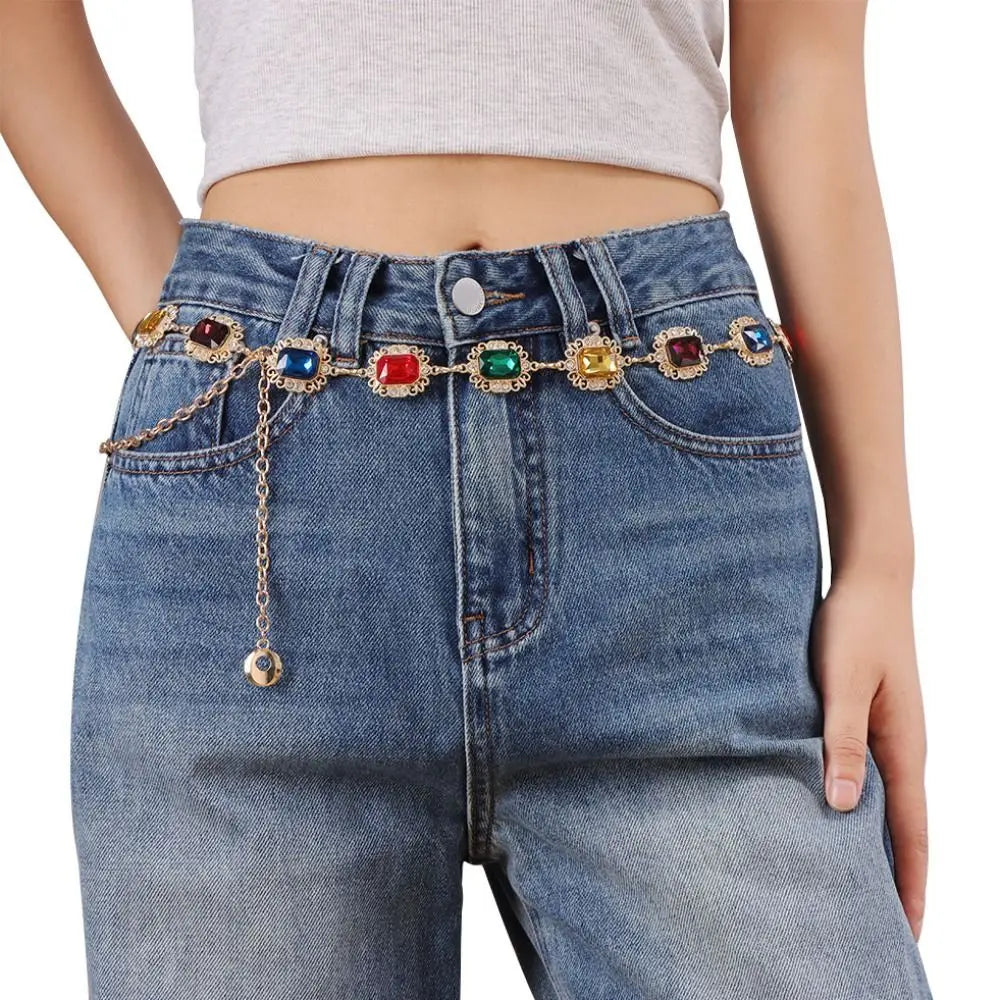A close-up of blue denim jeans featuring a multi-colored crystal waist chain embellishment.