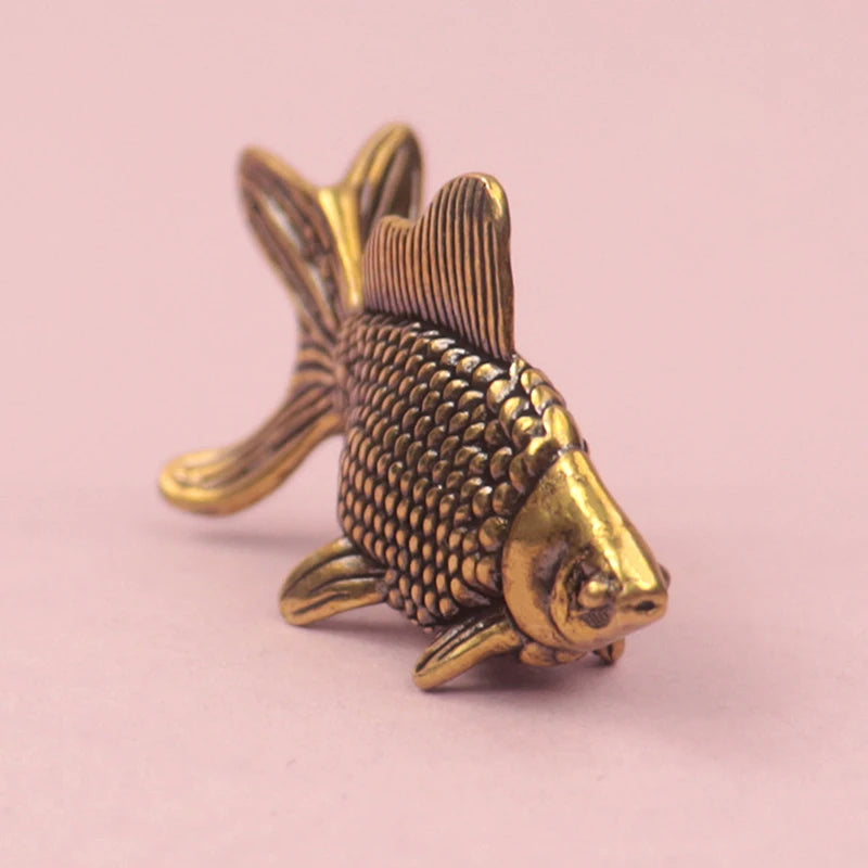 A small, detailed retro brass goldfish statue with textured scales and fins, resting on a pink surface.