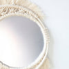 A round macrame wall mirror with fringe detailing in a natural beige color.