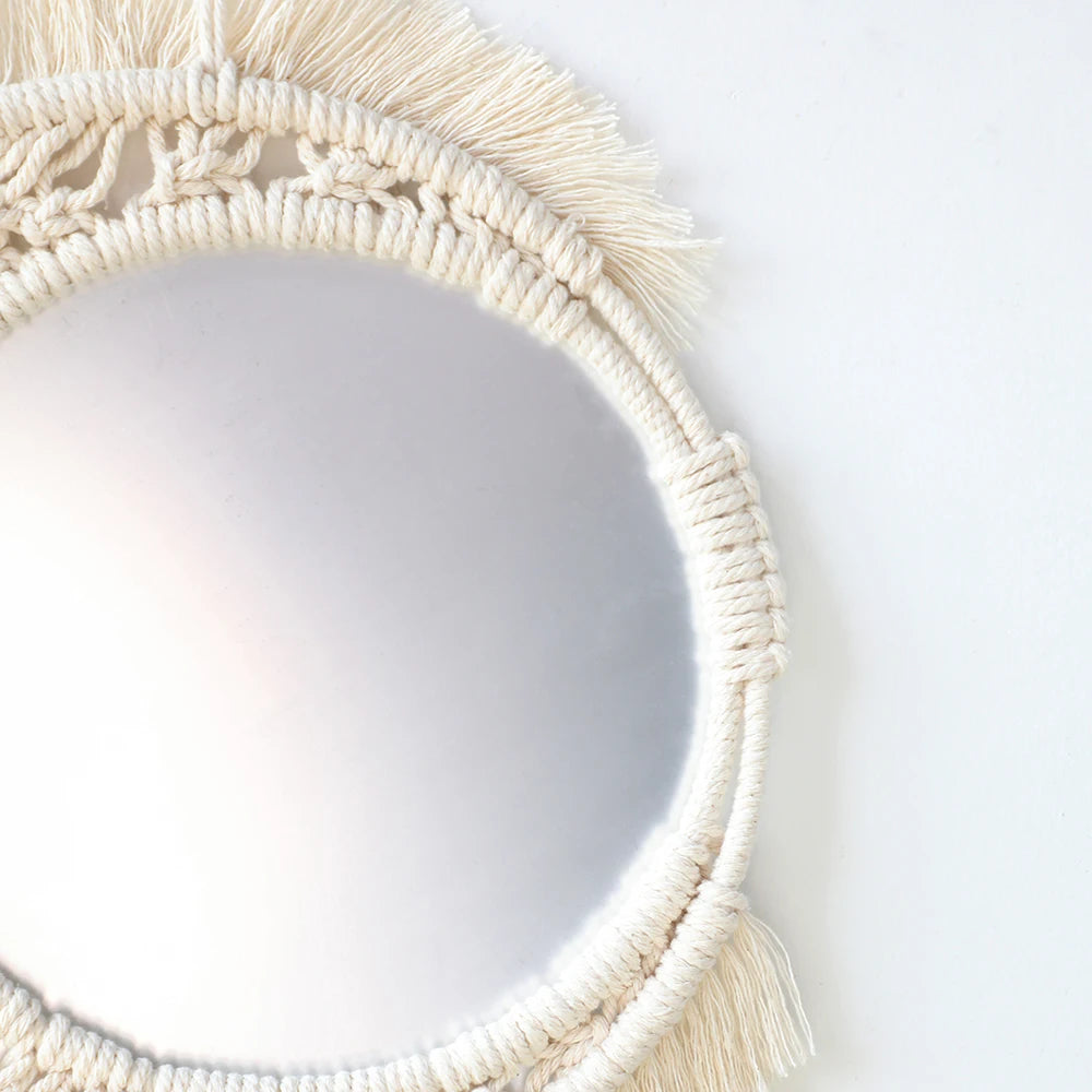 A round macrame wall mirror with fringe detailing in a natural beige color.