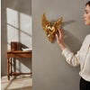 A woman's hand is placing an ornate golden winged heart wall decor on a gray wall.