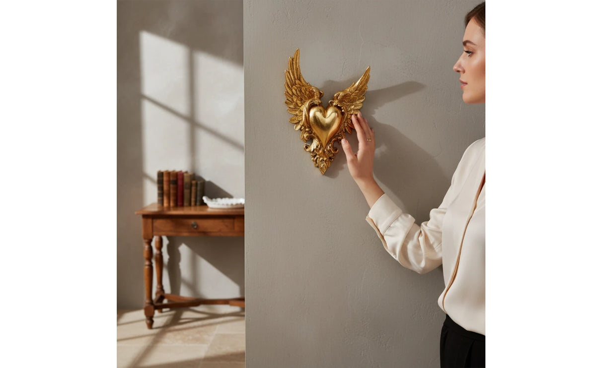 A woman's hand is placing an ornate golden winged heart wall decor on a gray wall.