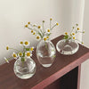A set of three clear glass bud vases holding small white and yellow wildflowers on a dark wooden surface.