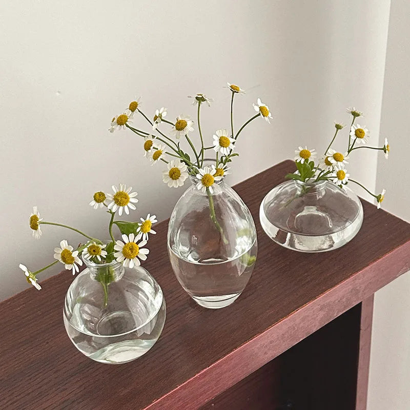A set of three clear glass bud vases holding small white and yellow wildflowers on a dark wooden surface.