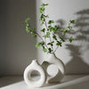 A white ceramic vase with a circular opening holds a sprig of green foliage against a light gray background.