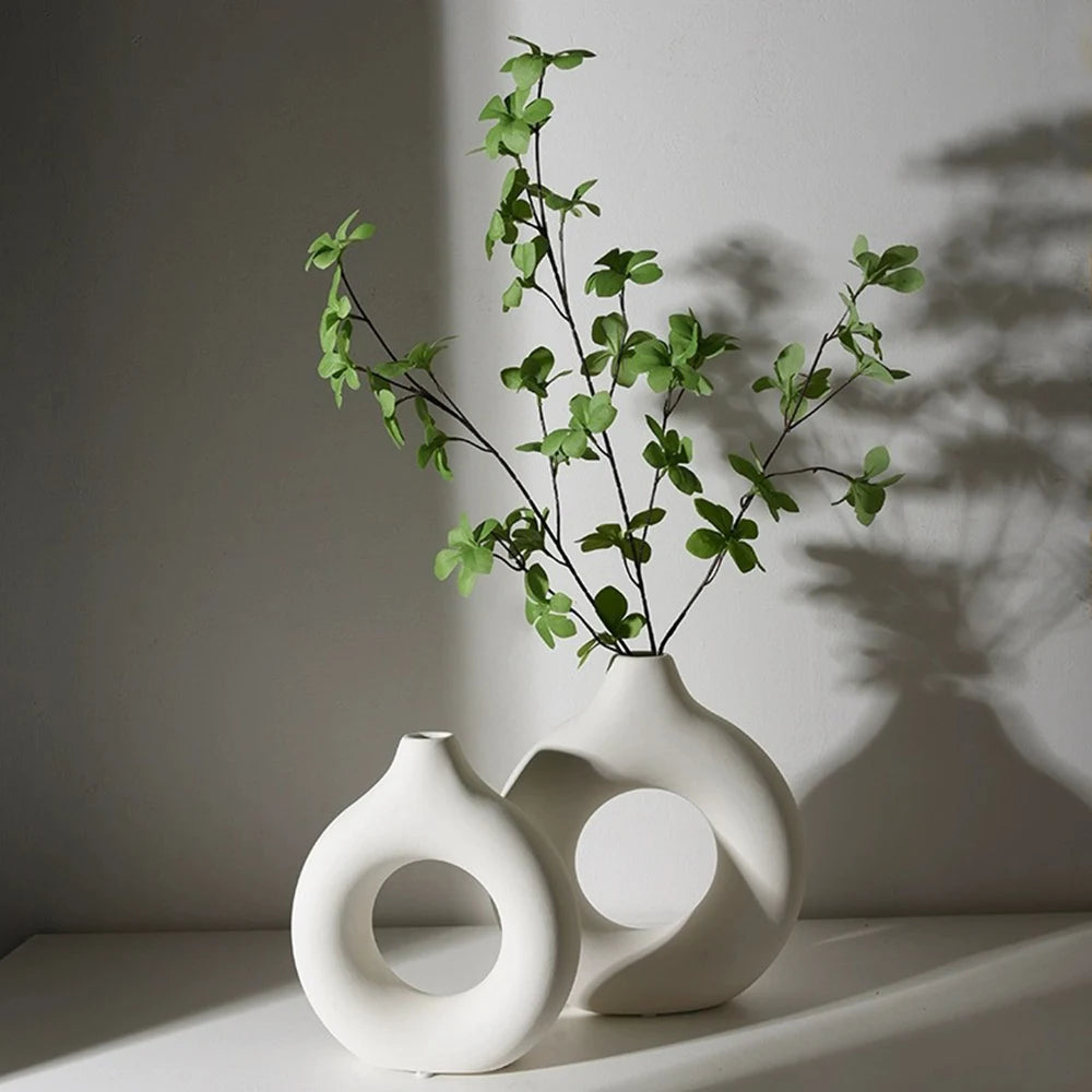 A white ceramic vase with a circular opening holds a sprig of green foliage against a light gray background.