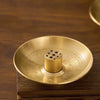 A round brass incense stick holder plate with a central cylindrical holder resting on a dark wooden surface.