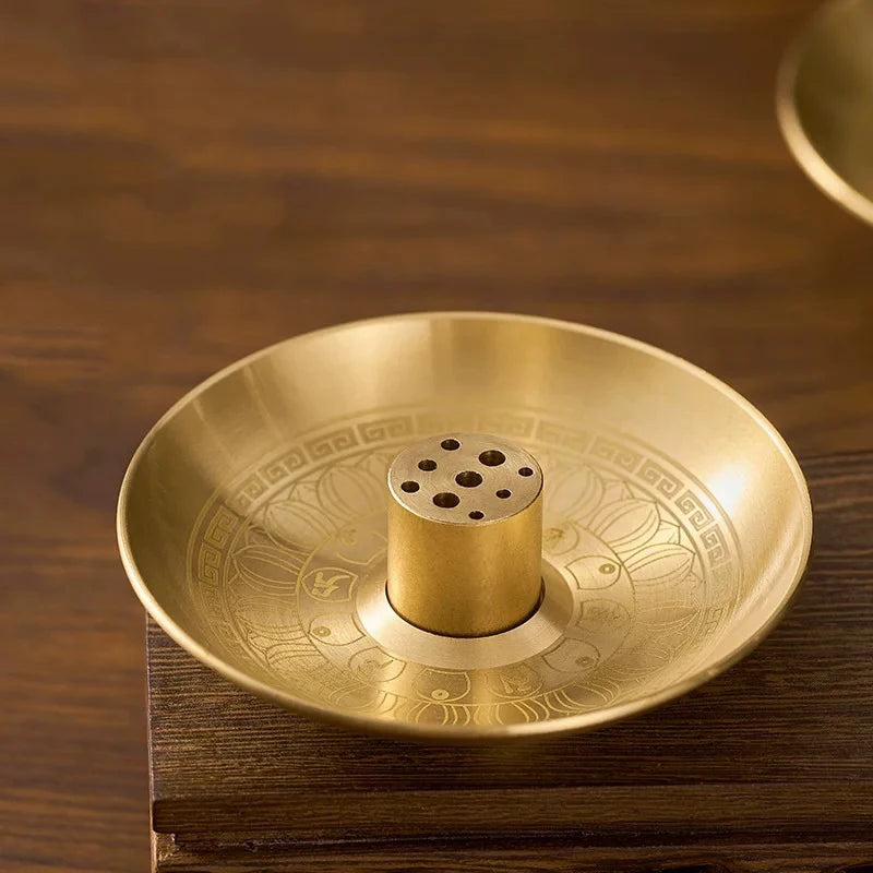 A round brass incense stick holder plate with a central cylindrical holder resting on a dark wooden surface.