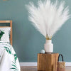A large bouquet of white artificial pampas grass stems in a light pink vase on a wooden nightstand.