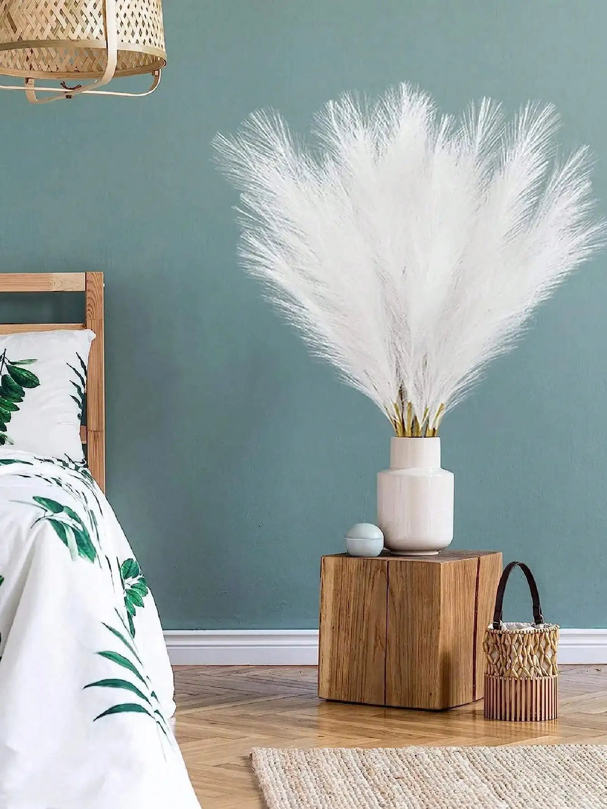 A large bouquet of white artificial pampas grass stems in a light pink vase on a wooden nightstand.