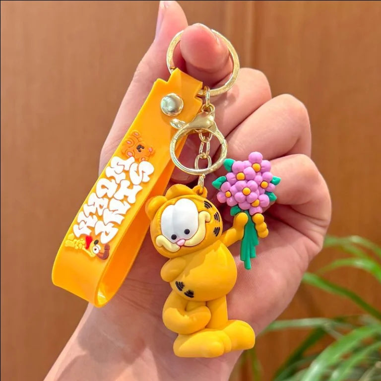 A hand holds a yellow Garfield cartoon keychain pendant with a gold ring and a purple flower charm.