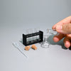 A hand holds a miniature laboratory set containing glass tubes and a small flask on a gray surface.