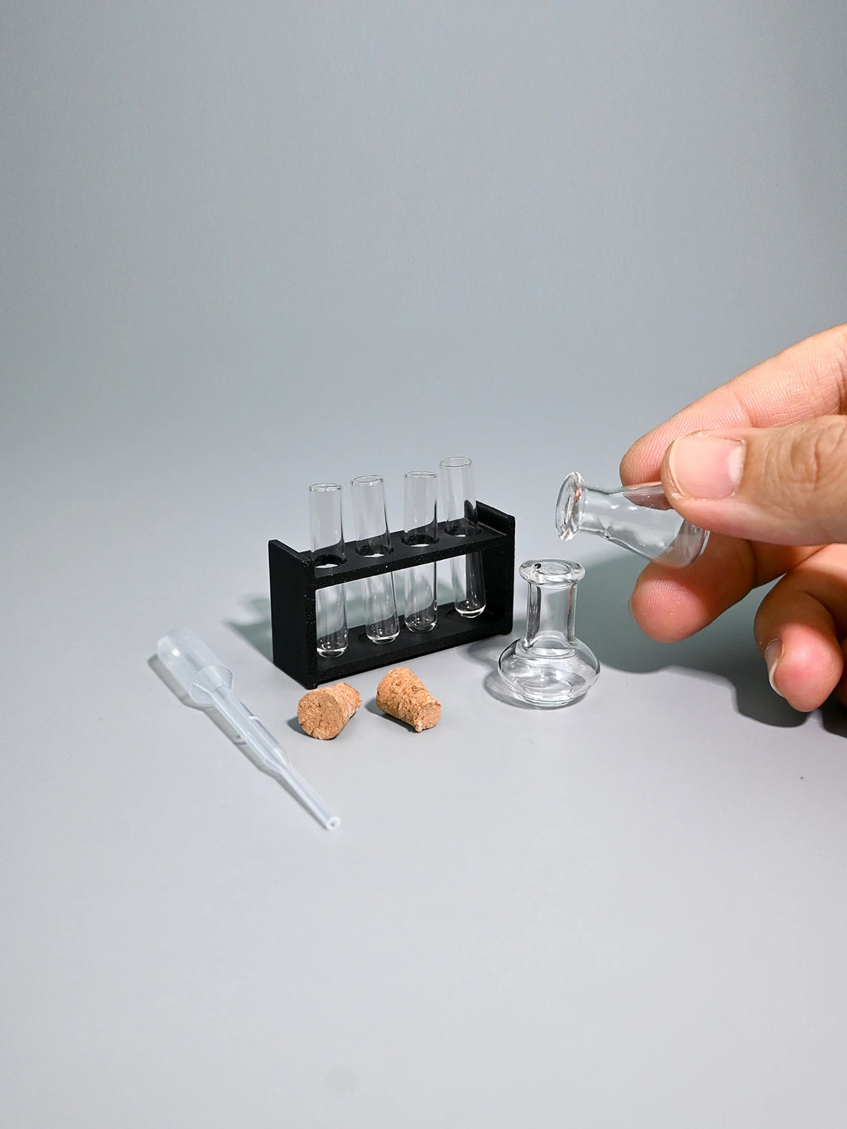A hand holds a miniature laboratory set containing glass tubes and a small flask on a gray surface.