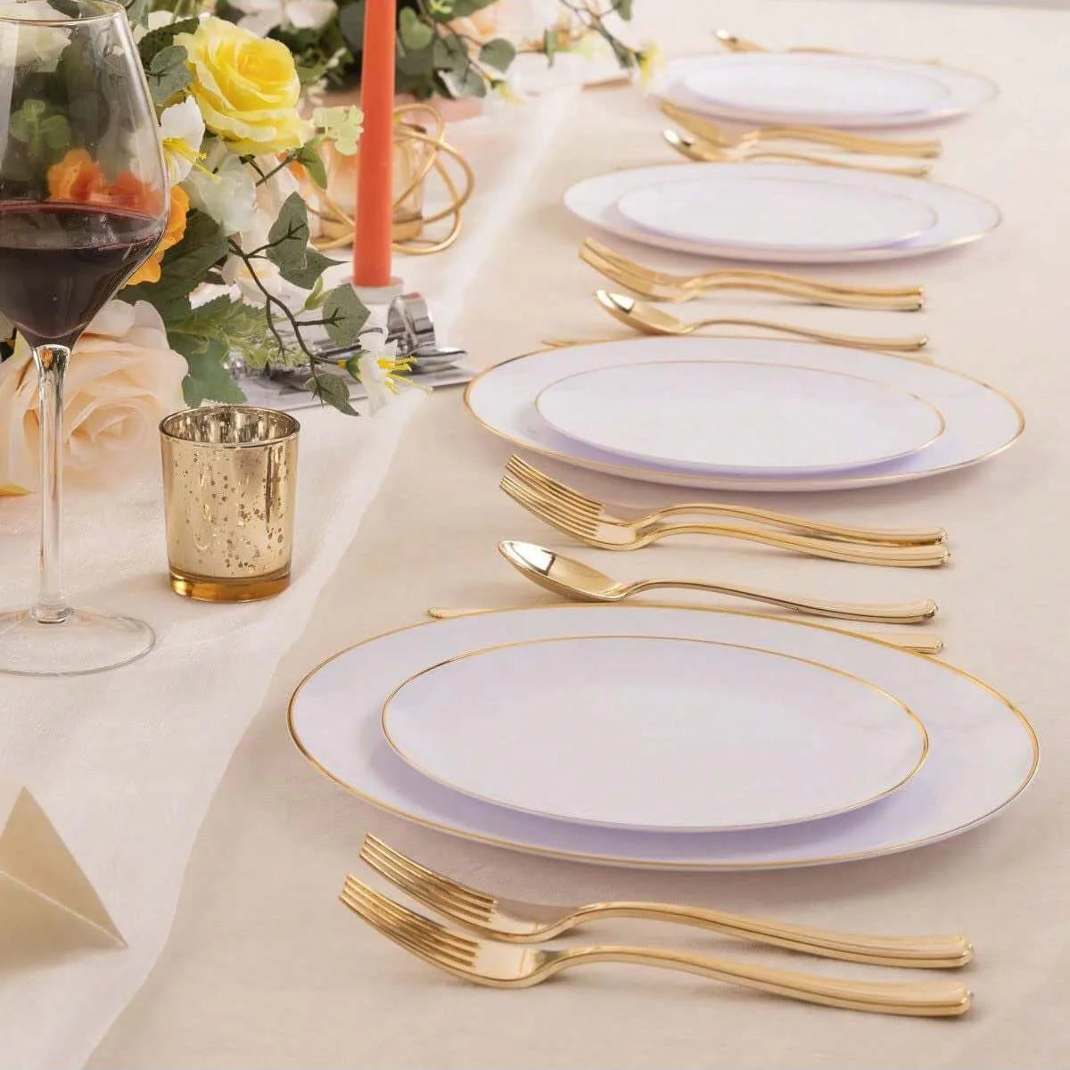A set of gold rim plastic plates for weddings and parties is displayed on a table with a glass of red wine and floral greener