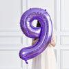 A purple foil number balloon shaped like the number 9 is held by a person against a white paneled background.