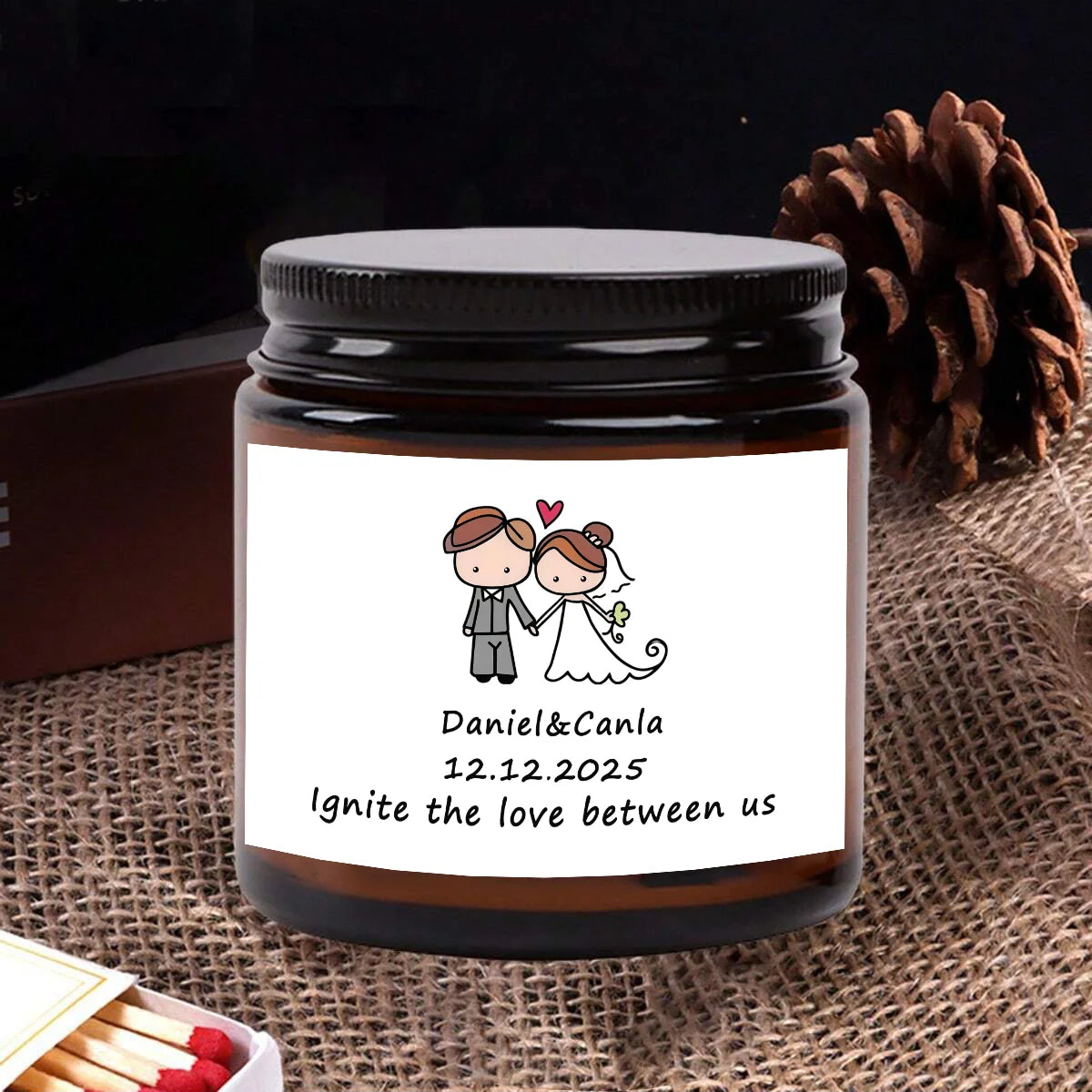 A dark glass jar candle with a label featuring the names Daniel & Canla and the date 12.12.2025