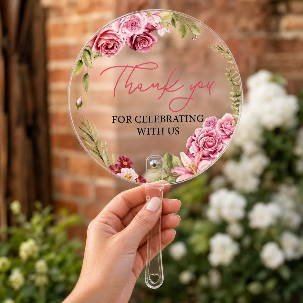 A hand holds a clear glass thank you sign with pink floral designs and greenery.