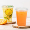 Two clear plastic drinking cups filled with orange juice and garnished with citrus slices resting on a wooden surface.