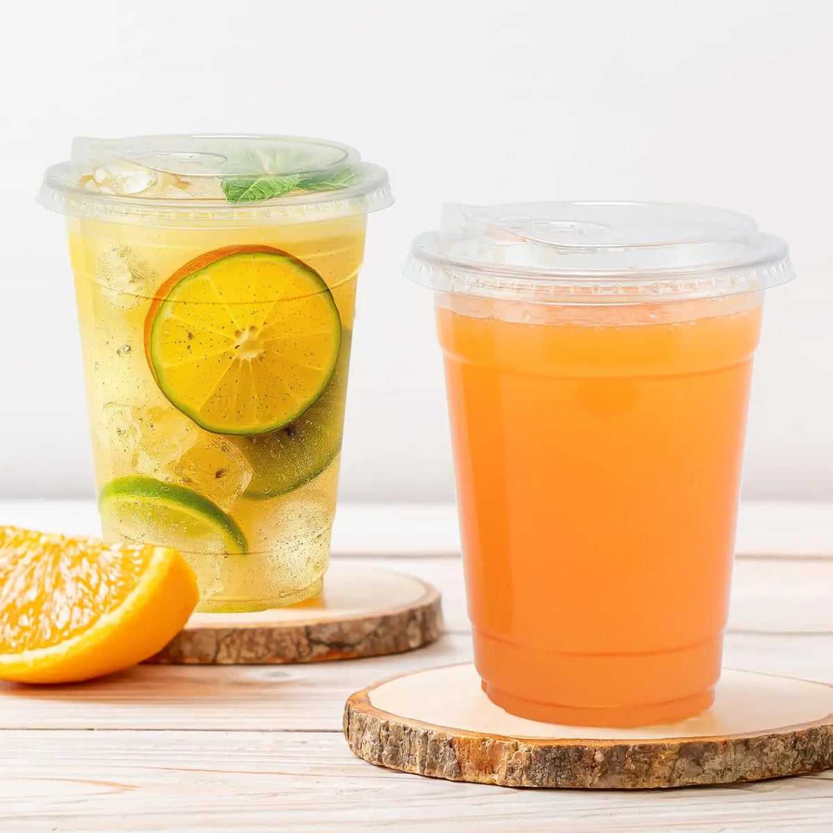 Two clear plastic drinking cups filled with orange juice and garnished with citrus slices resting on a wooden surface.