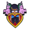 A enamel pin featuring a cartoon character with a black hair, a green unicorn, and a pink unicorn surrounding a heart shape.