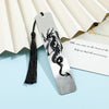 A silver metal bookmark featuring a black dragon design with a tassel, resting on a light blue surface.