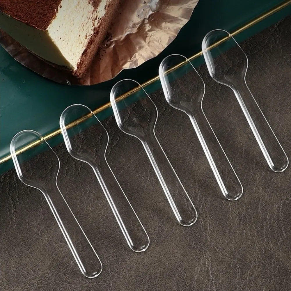 Five clear plastic disposable spoons arranged diagonally on a dark surface next to a slice of cake.