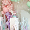 A pink and purple mermaid cake topper set featuring sea elements like shells, seaweed, and colorful balloons.