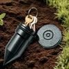 A dark metal key hider attached to a watering can, resting on brown soil with green foliage.