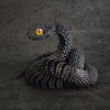 A dark, textured 3D printed snake art model with an amber eye, resting on a dark gray surface.