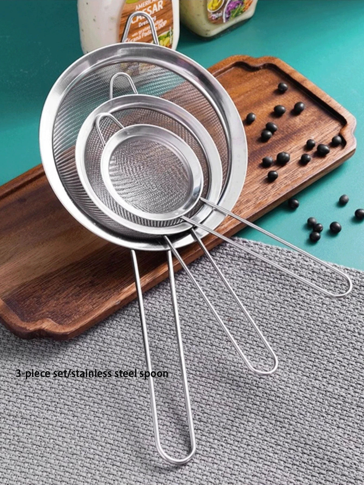 A set of three stainless steel colanders and strainers resting on a wooden board and gray fabric.