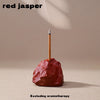 A stick of red jasper is shown resting on a reddish-brown rock, with smoke rising above it.