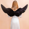 A woman wearing a white dress and black angel feather wings against a beige background.