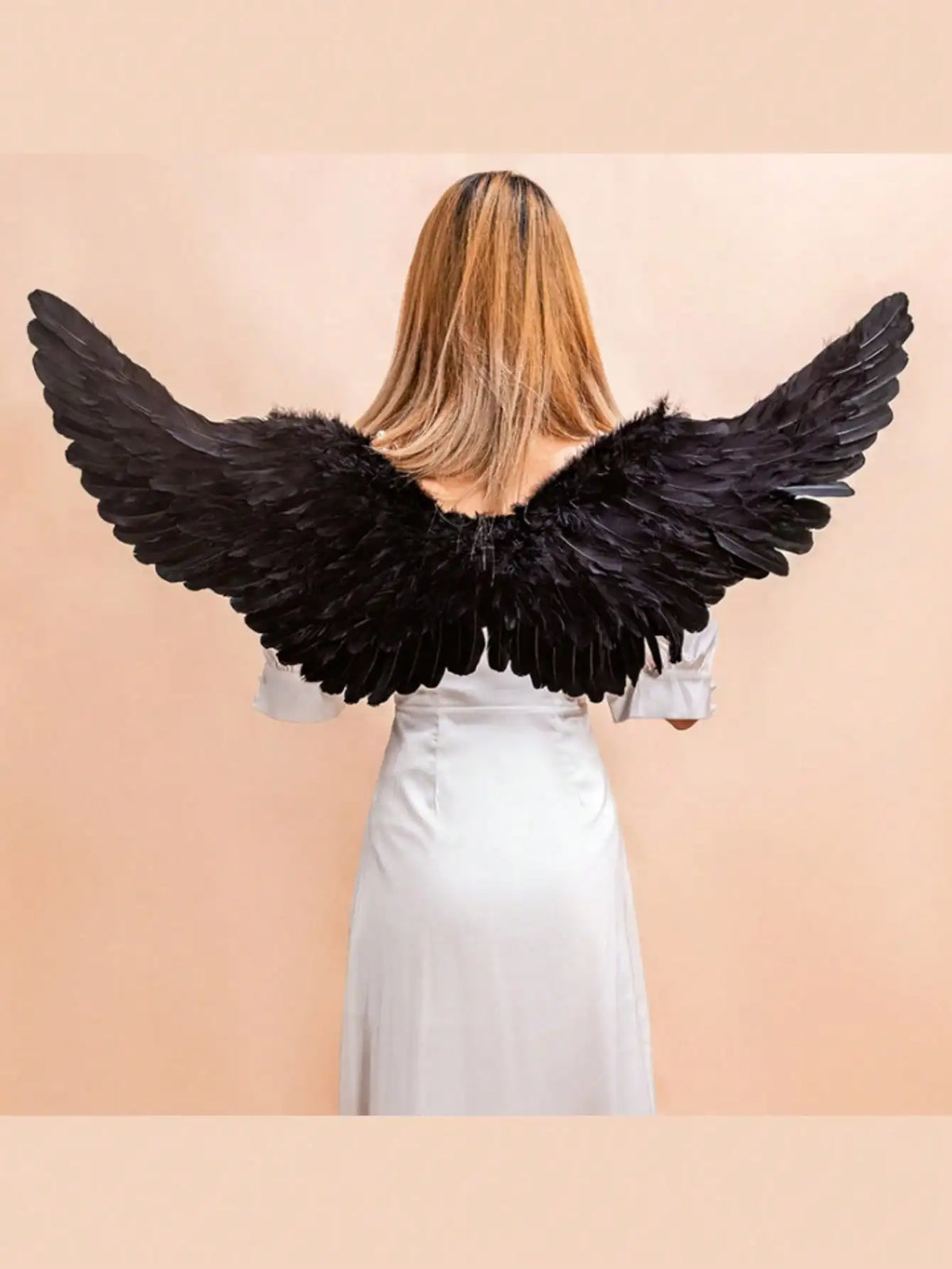 A woman wearing a white dress and black angel feather wings against a beige background.