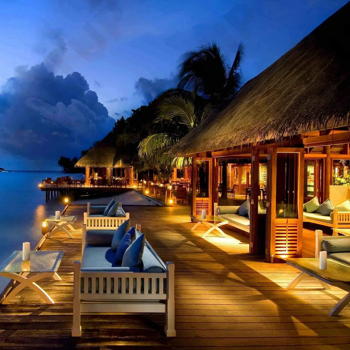 A tropical wooden deck at dusk featuring comfortable outdoor seating and illuminated by warm lighting.