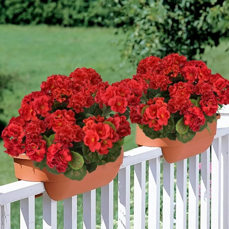 A terracotta planter filled with vibrant red faux silk geranium flowers displayed on a white picket fence.