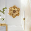 Gold flower-shaped resin wall art mounted on a white wall next to a sheer white curtain and a gold frame.