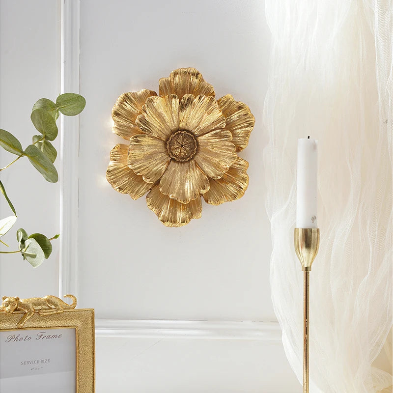 Gold flower-shaped resin wall art mounted on a white wall next to a sheer white curtain and a gold frame.